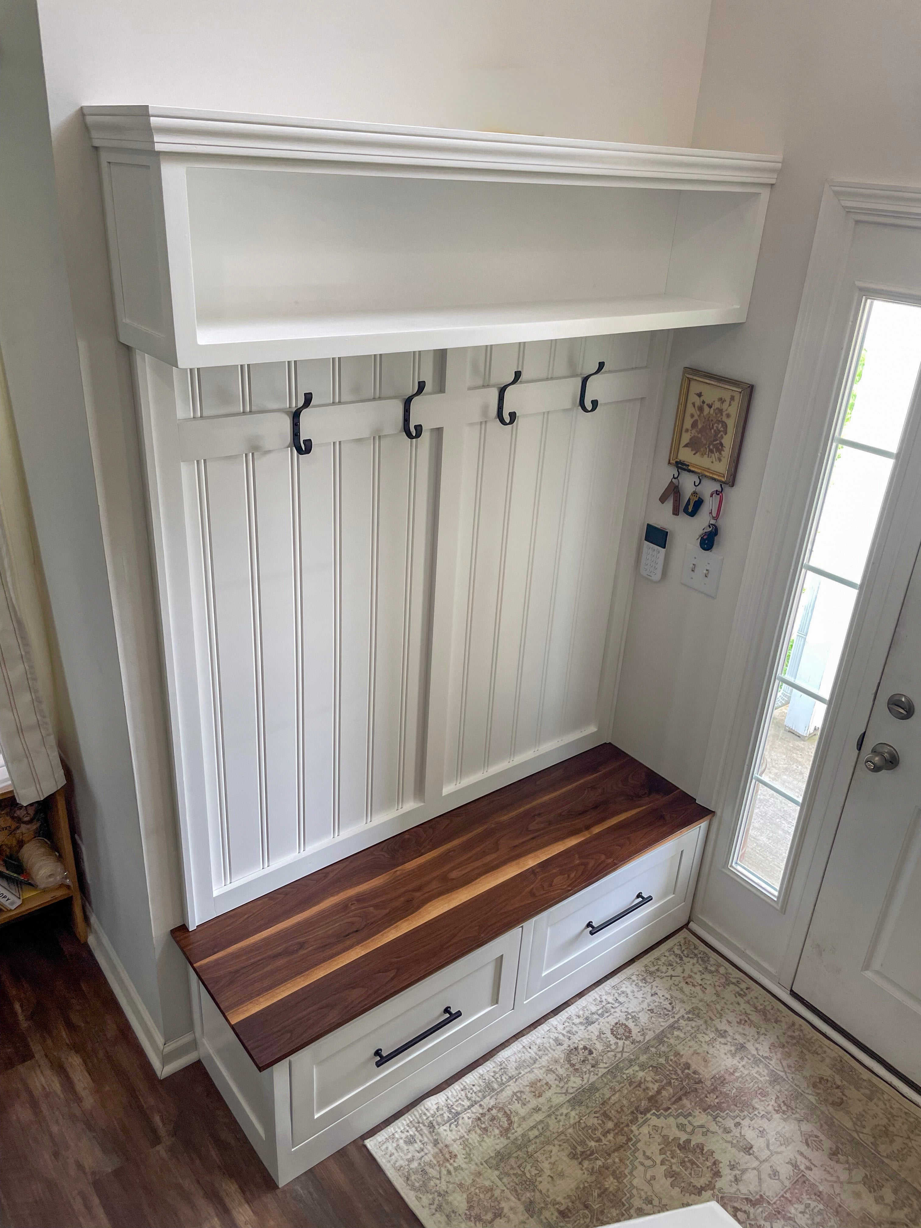 A white entryway mudroom bench with a walnut bench, built-in drawers, black coat hooks, and an overhead shelf, shown in a home entryway.