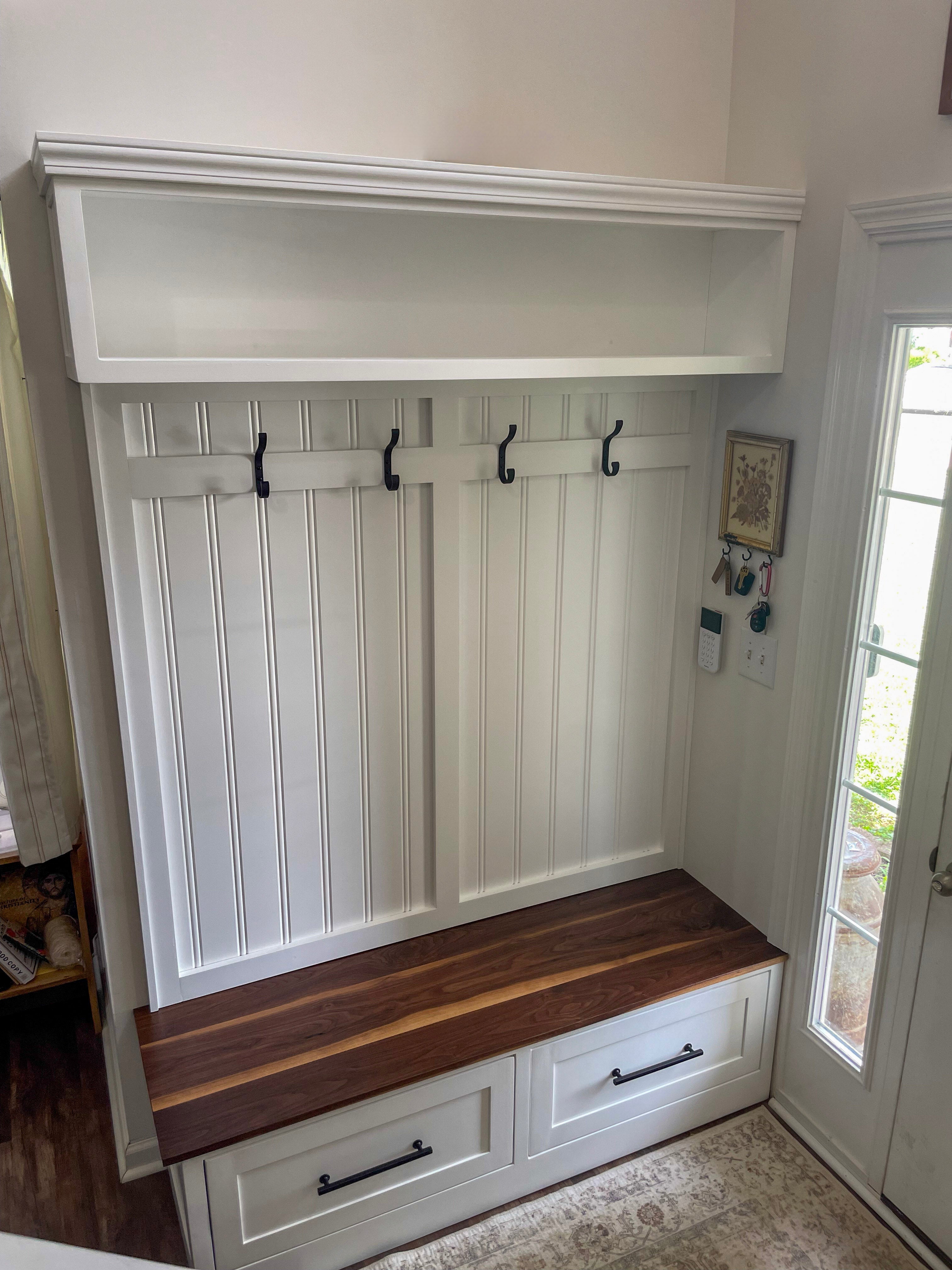 A white entryway mudroom wall unit with a bench, four black coat hooks, and an open shelf, shown in a real interior next to a glass door.