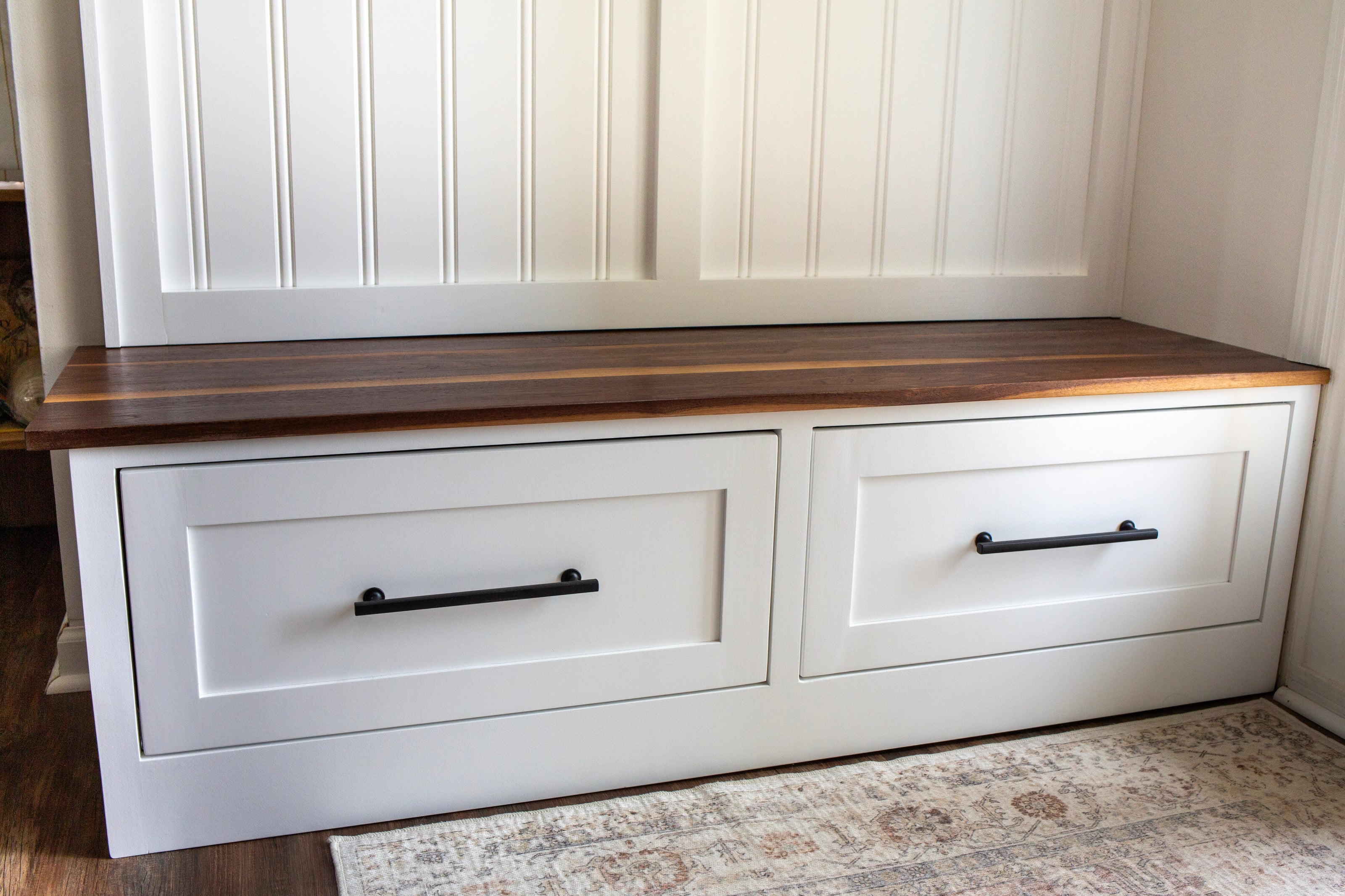 A white wooden storage bench with two drawers and a walnut top, set against a white paneled wall with black metal drawer pulls.
