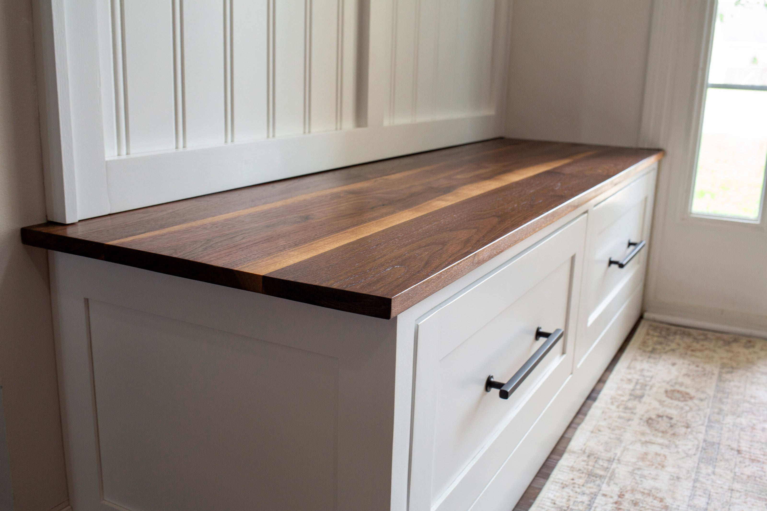 A white built-in storage bench with a thick, dual-toned dark wood countertop and black metal drawer pulls, set in a bright room with white paneled walls and a window.